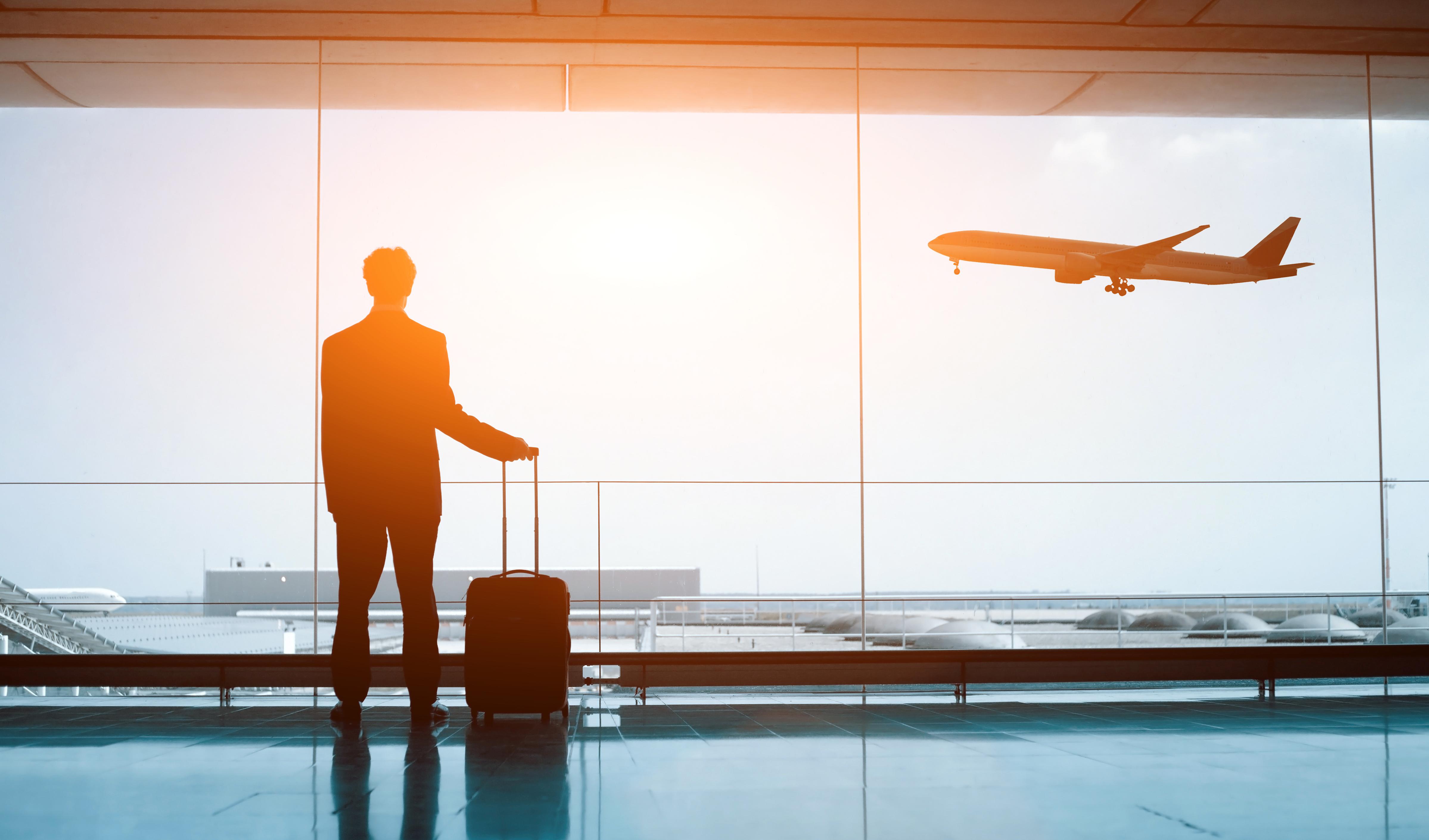 Gesund reisen Mann steht am Flughafen mit einem Koffer und schaut einem Flugzeug bei der Landung zu